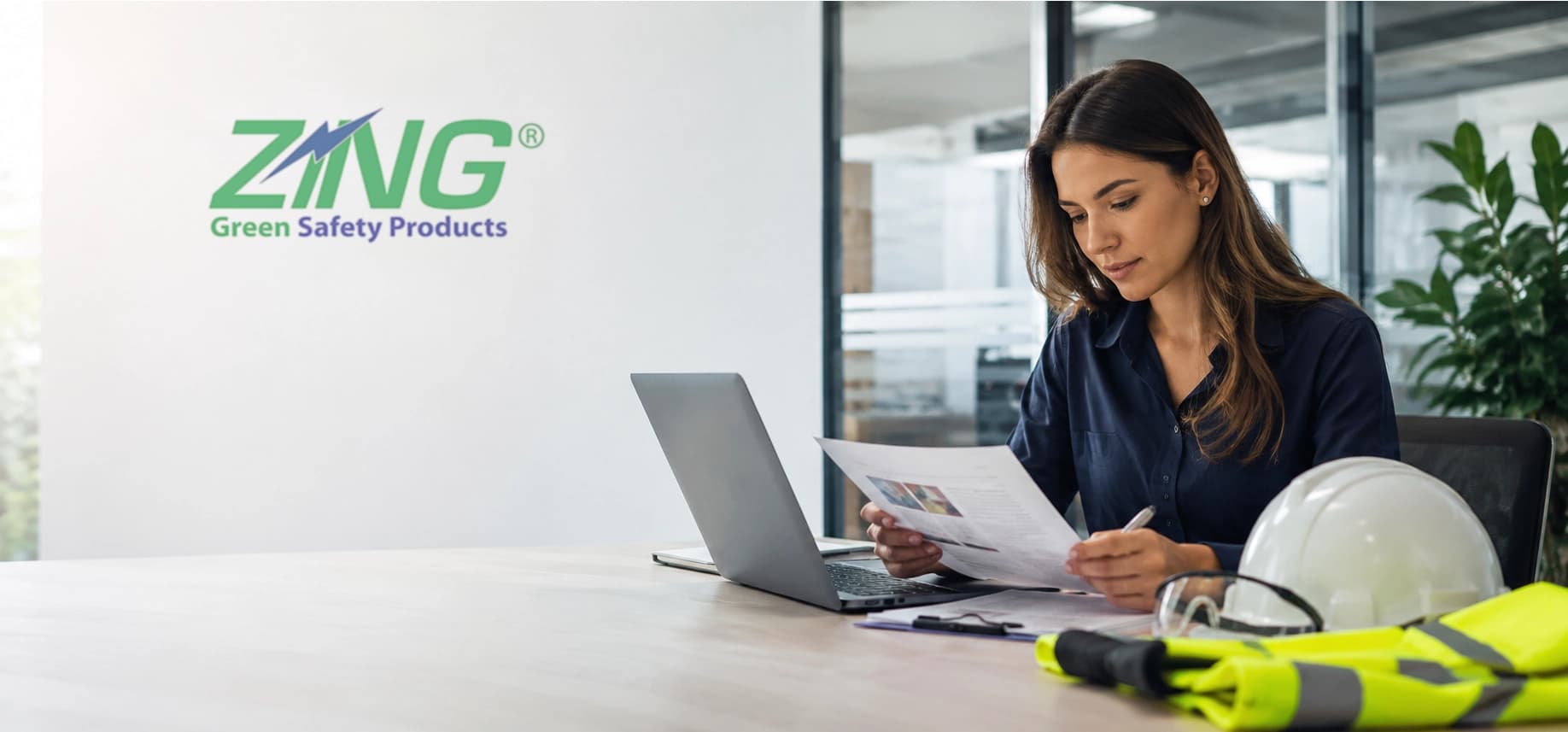 A woman works at a desk with a laptop, documents, and safety gear. The wall displays "ZING Green Safety Products" signage.